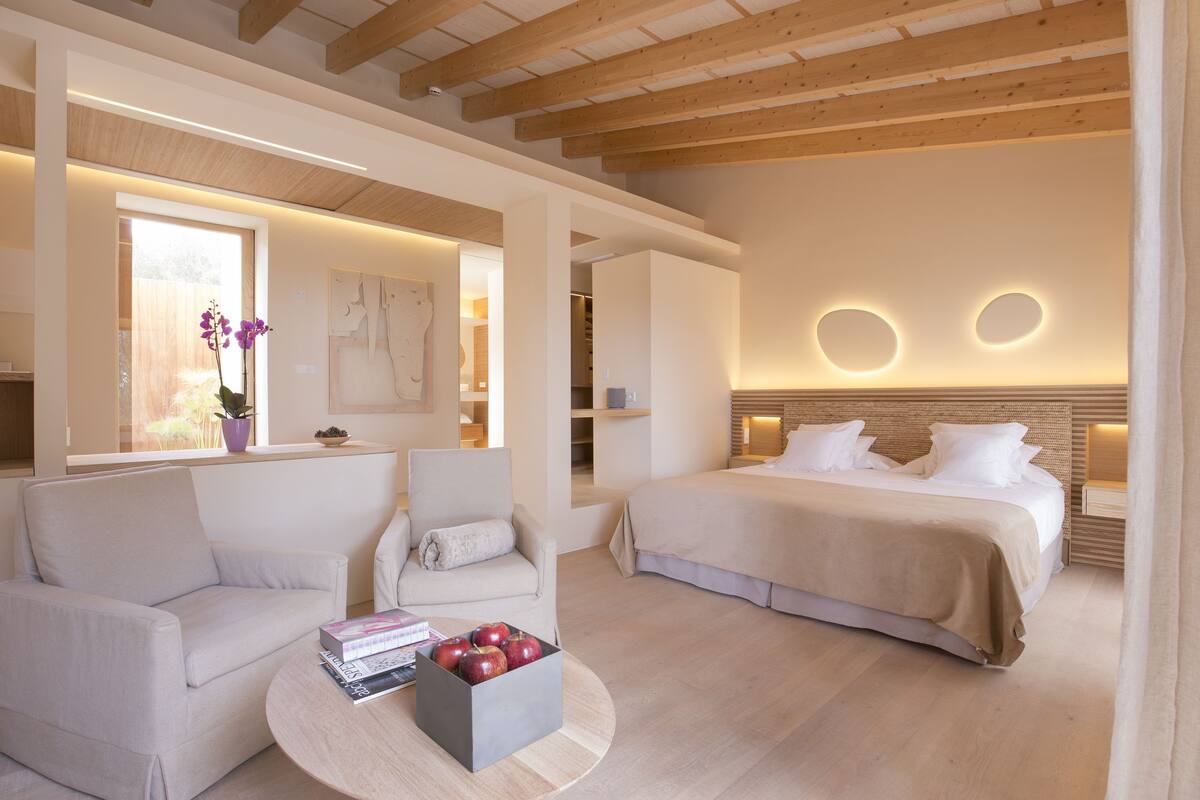 Une chambre d’hôtel moderne avec un grand lit, un canapé, une table basse avec des livres et des pommes, et un plafond en bois.