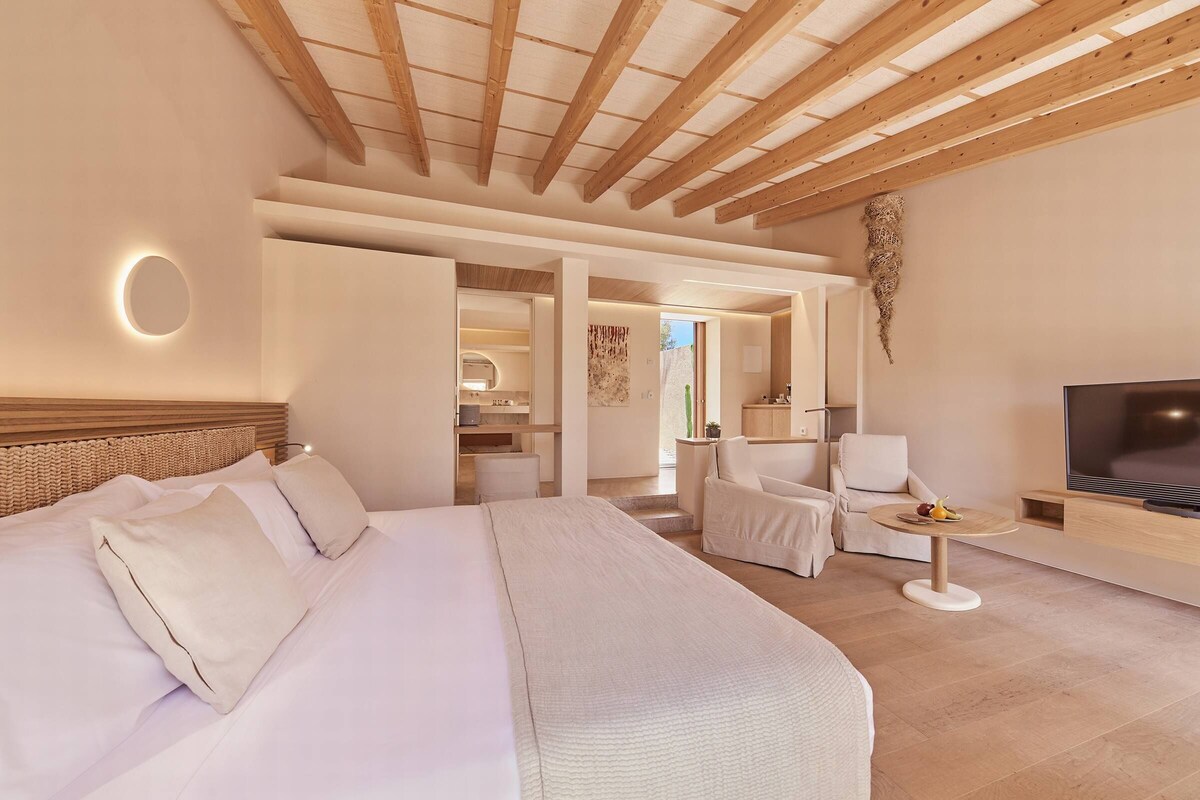 Una habitación de hotel moderna con una cama grande, televisor, zona de estar y baño visible a través de una puerta abierta.