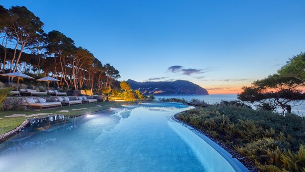 Une piscine entourée d’arbres, avec des chaises longues et des parasols, offrant une vue sur la mer au coucher du soleil.