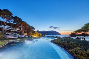 A swimming pool with lounge chairs and umbrellas, surrounded by trees and a view of the sea at sunset.