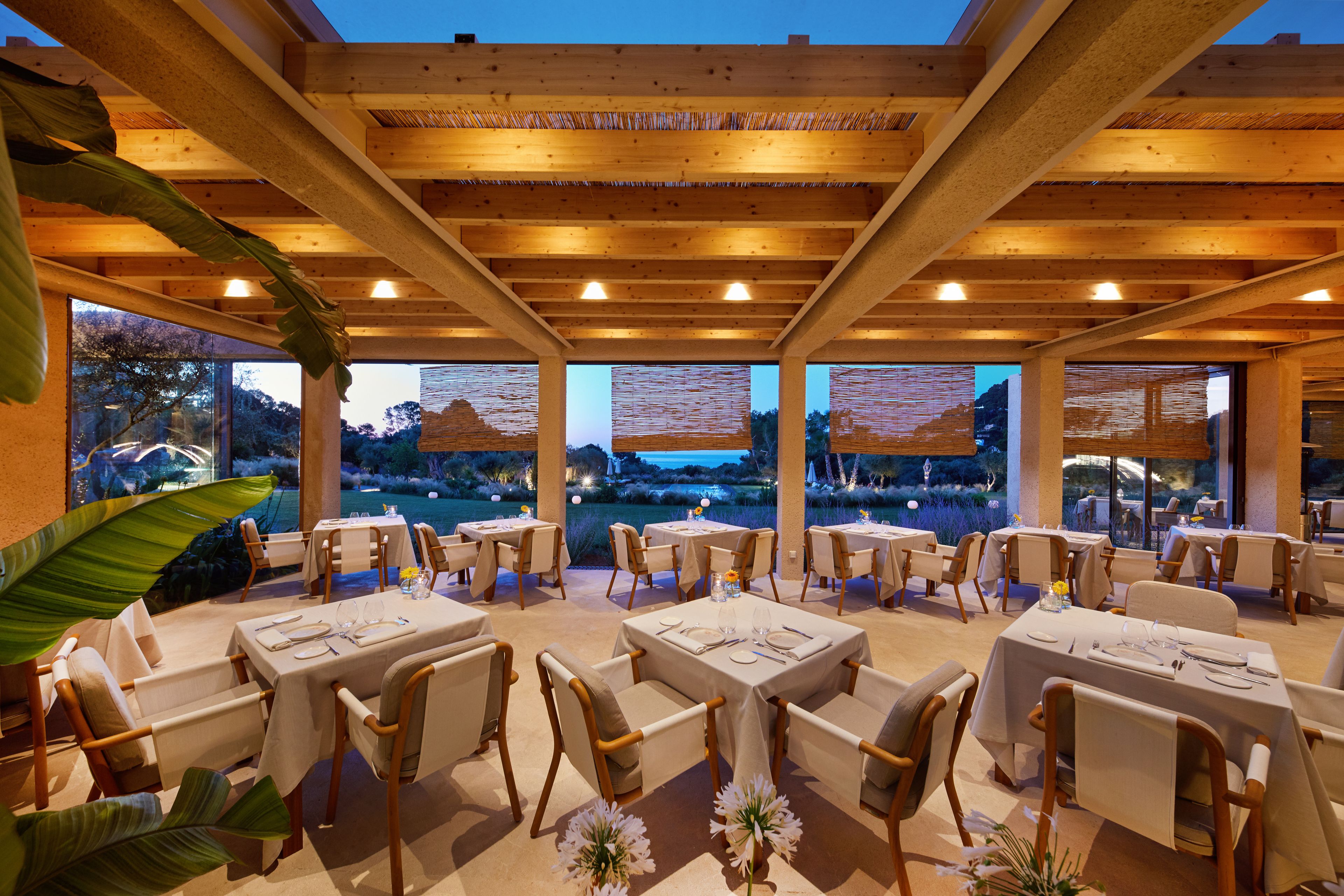 A restaurant with wooden beams, tables set for dining, and large windows overlooking a scenic view.