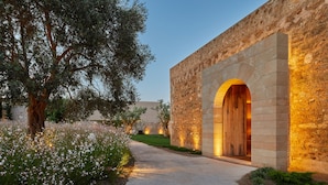 Un bâtiment en pierre doté d’une entrée en arc de cercle, un chemin menant à celui-ci et un jardin agrémenté de plantes à fleurs.