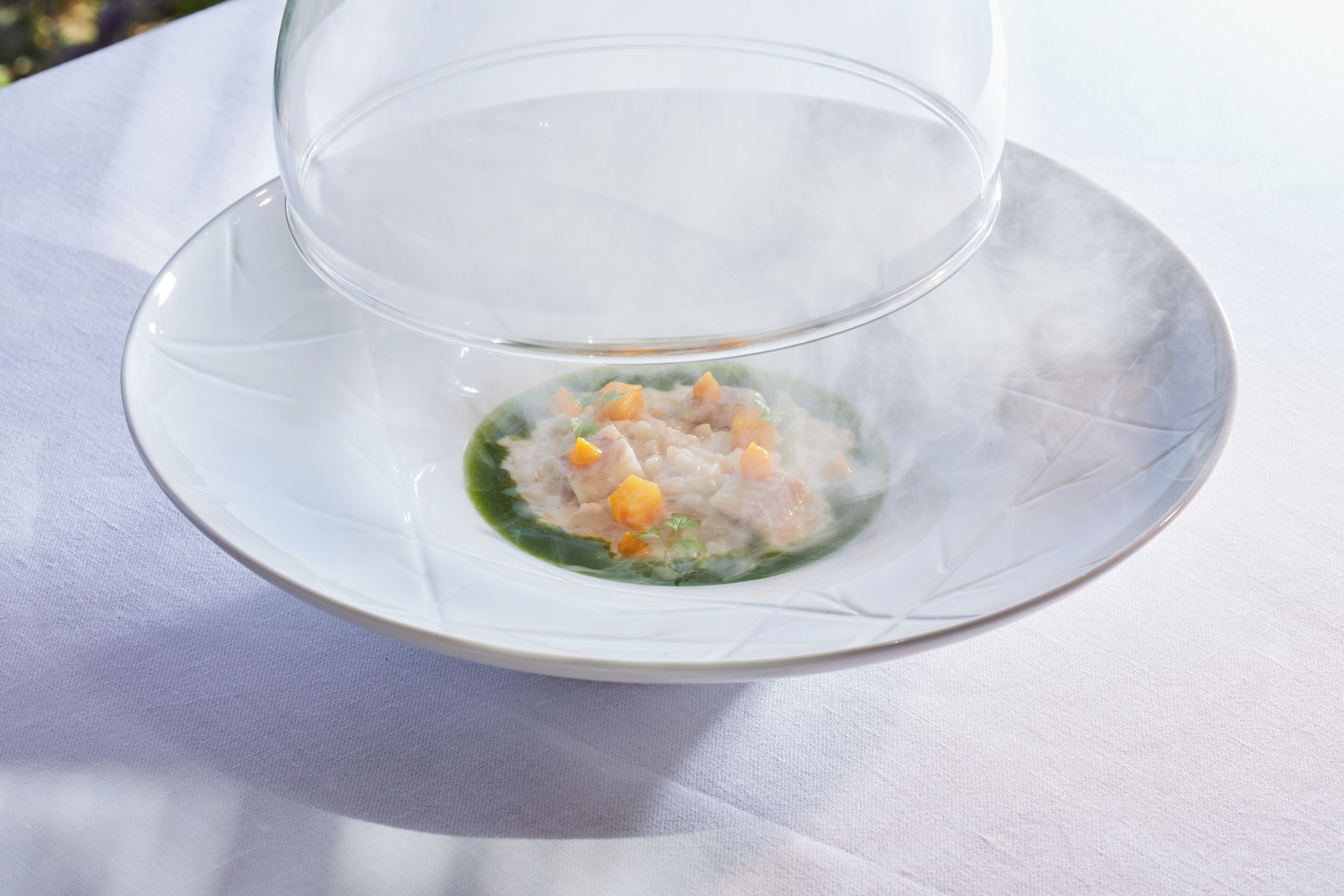Un tazón de vidrio transparente con salsa verde e ingredientes picados sobre un mantel blanco.