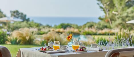 Una mesa preparada para el desayuno con vista al océano, rodeada de follaje.