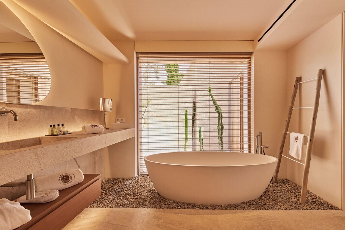 Une salle de bain moderne avec une baignoire sur pieds, un meuble-lavabo en bois, un grand miroir et une fenêtre avec des stores.