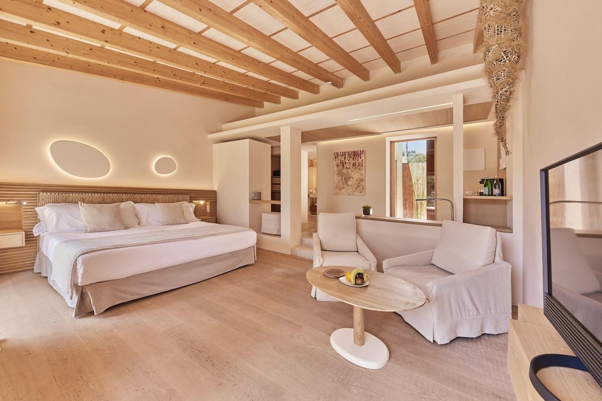 Quarto de hotel moderno com teto de madeira, uma cama grande, um sofá e uma pequena mesa com frutas.