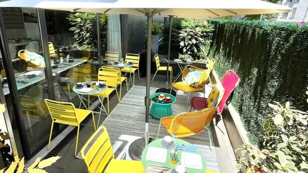 Un espace extérieur aménagé avec des chaises et des tables jaunes, protégé par un parasol blanc, et une haie verte le long du bâtiment.