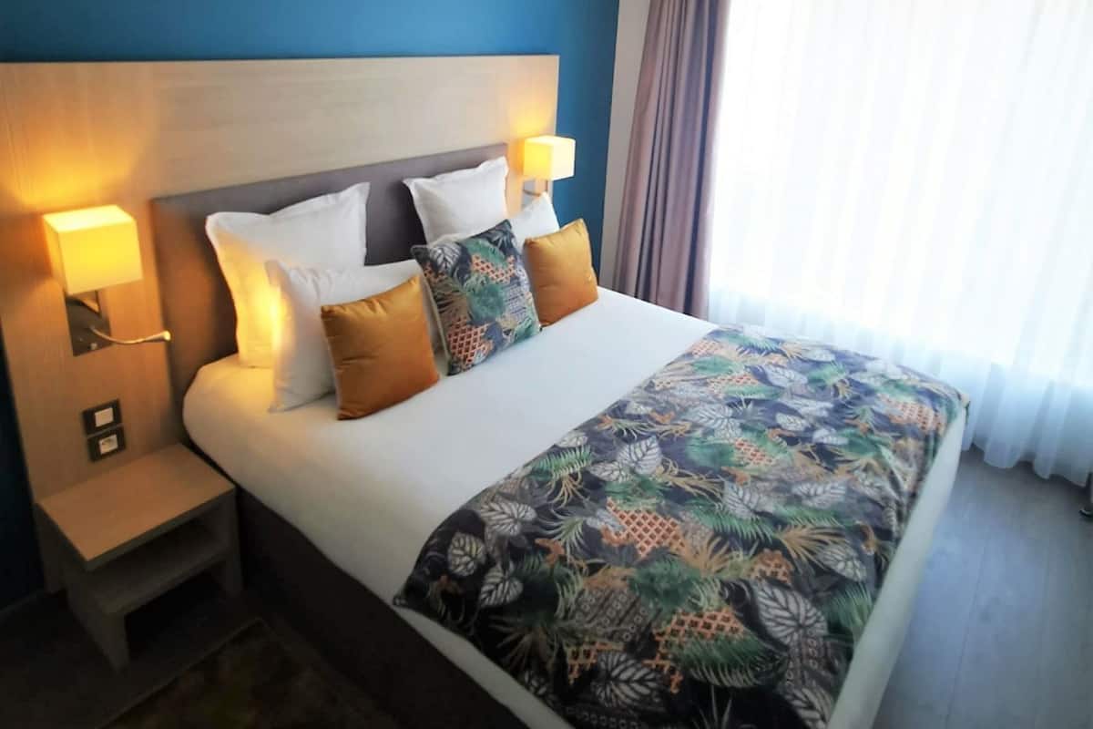 Una habitación de hotel con cama, mesitas de noche, cabecera y ventana con cortinas.
