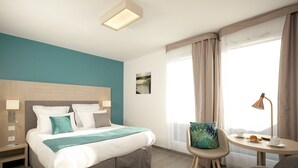 Une chambre d’hôtel comprenant un lit, des tables de chevet, une chaise, une petite table avec une lampe et une fenêtre avec des rideaux.
