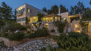 A modern two-story house with a stone facade, multiple balconies, and a well-lit garden area. - Electra Beachfront Villa (Ermionida)