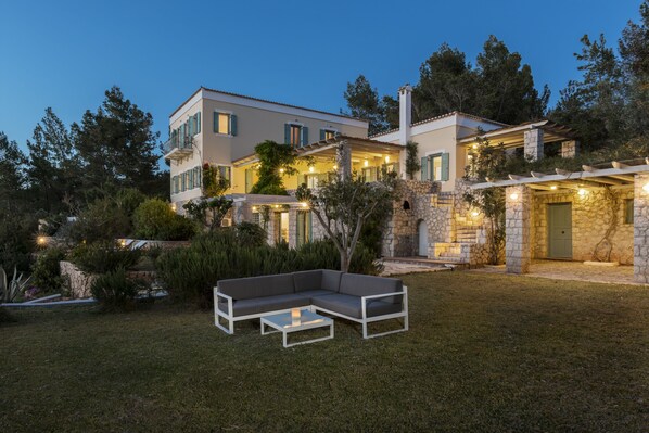 A modern two-story house with a stone exterior, a large outdoor seating area, and a well-lit entrance. - Electra Beachfront Villa (Ermionida)