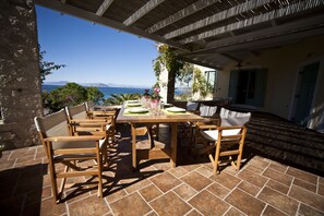 An outdoor dining area with a view of the sea, wooden furniture, and a stone wall. - Electra Beachfront Villa (Ermionida)
