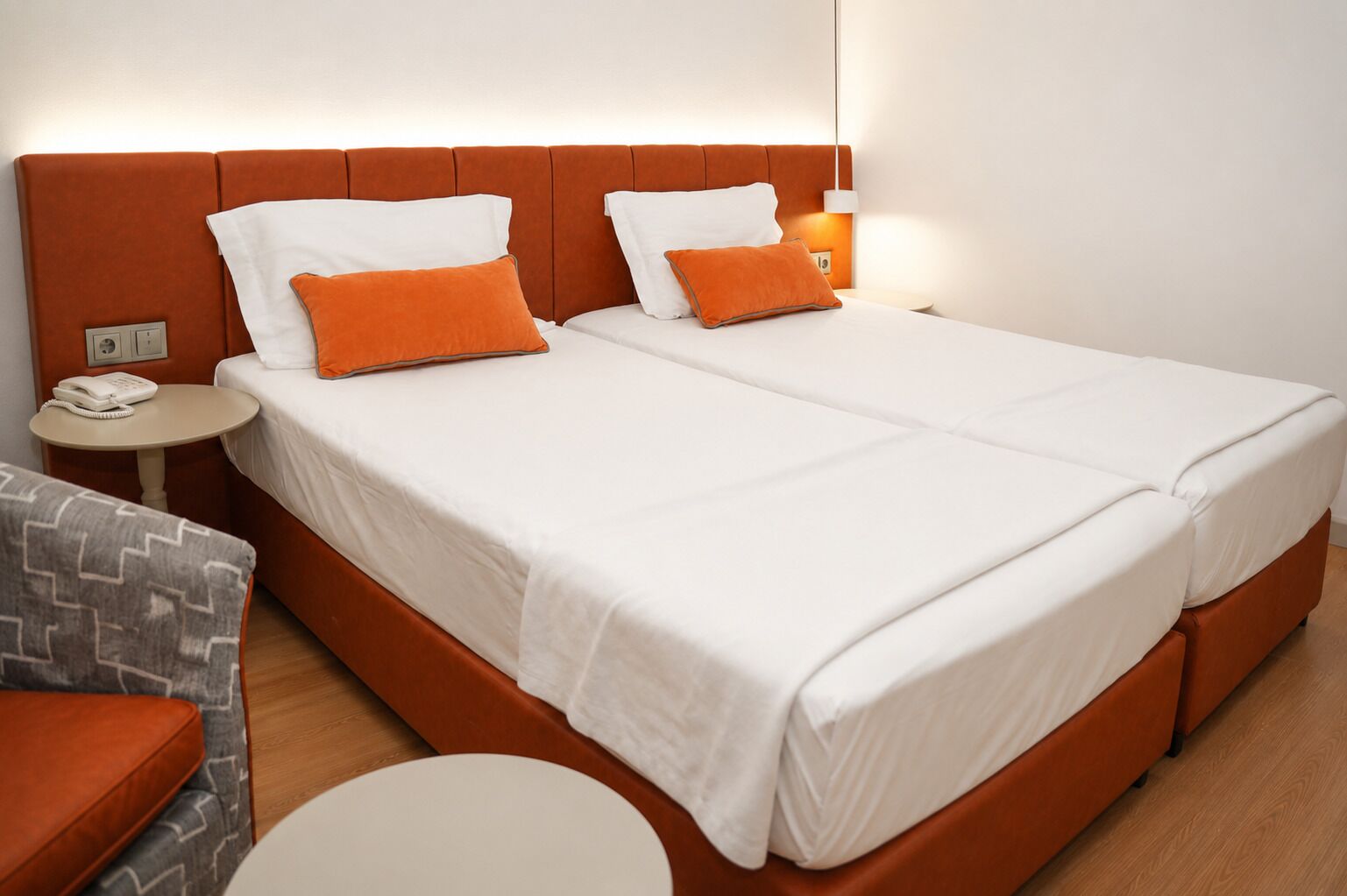 Cama doble con ropa de cama blanca y almohadas anaranjadas, una mesita de noche con teléfono y una pequeña mesa redonda.