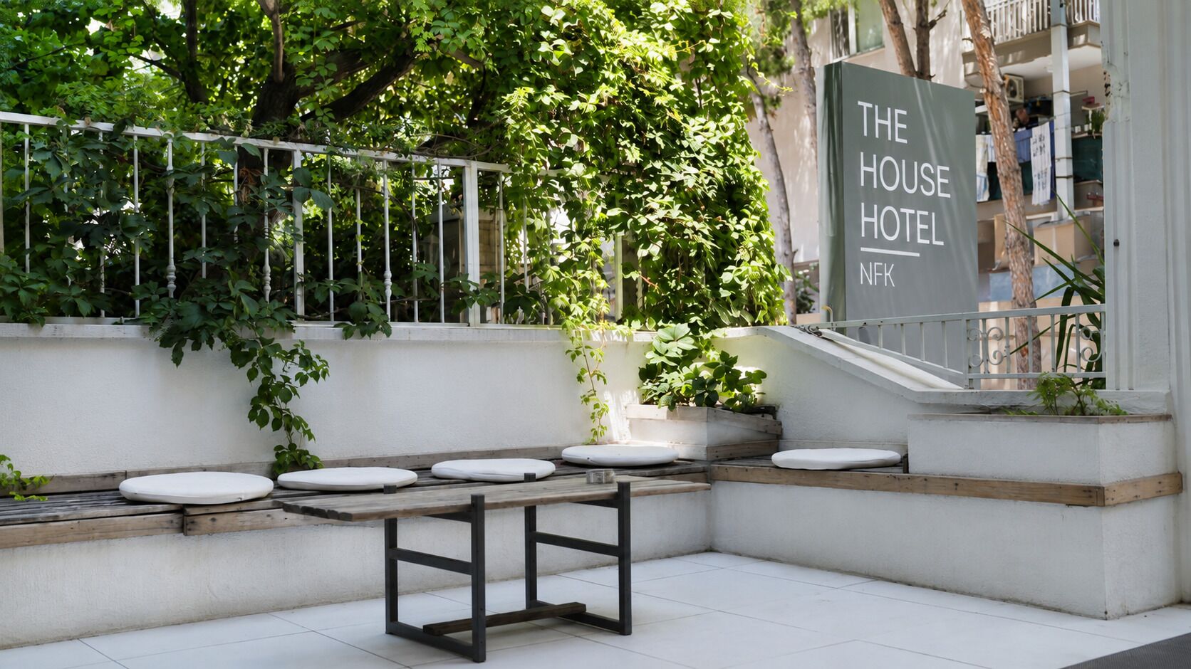 Un área de estar al aire libre con cojines blancos y una mesa de madera, rodeada de follaje y una escalera que lleva a un edificio con un letrero que dice 'THE HOUSE HOTEL NFK'.