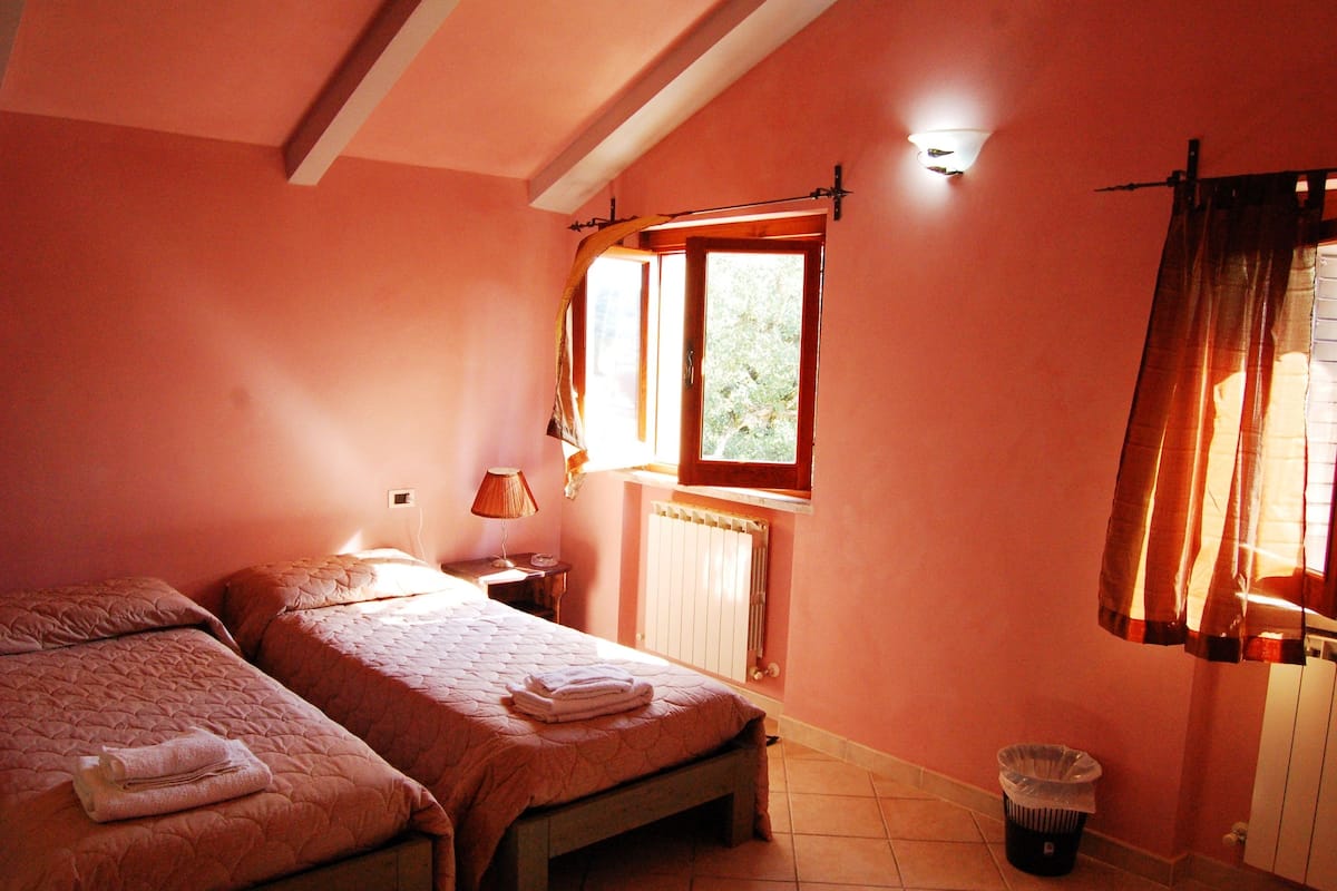 Quarto com duas camas, uma janela com cortinas, luminária e radiador.