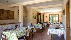 Un espace repas avec plusieurs tables recouvertes de nappes à motifs floraux, des chaises en bois et un mur de briques orné d’un grand tableau.