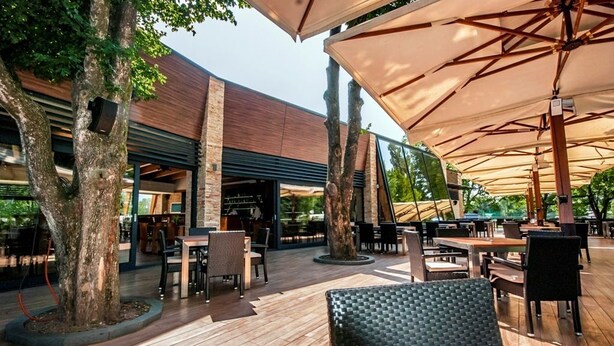 Un patio al aire libre con suelo de madera, mesas, sillas y grandes sombrillas que ofrecen sombra.