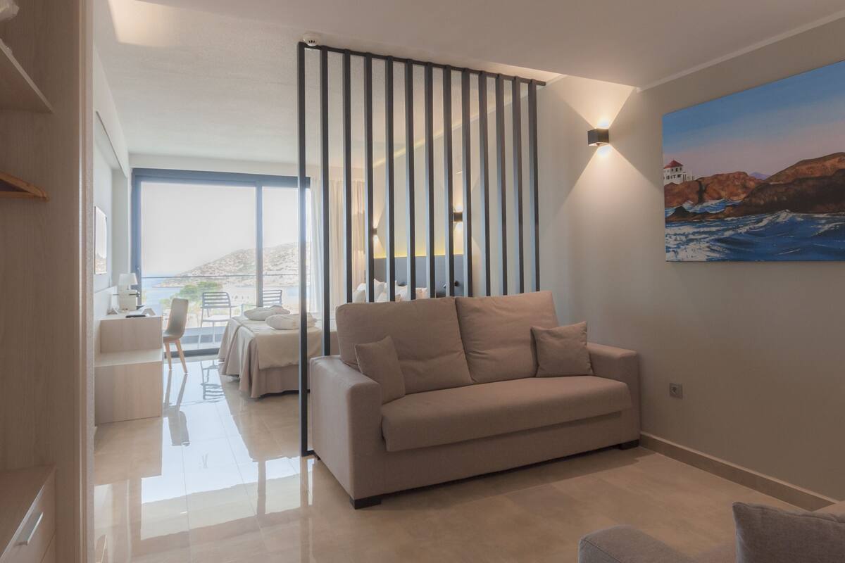 Una sala de estar moderna con un sofá color beige, un comedor con mesa y sillas, y un cuadro grande de un paisaje costero en la pared.