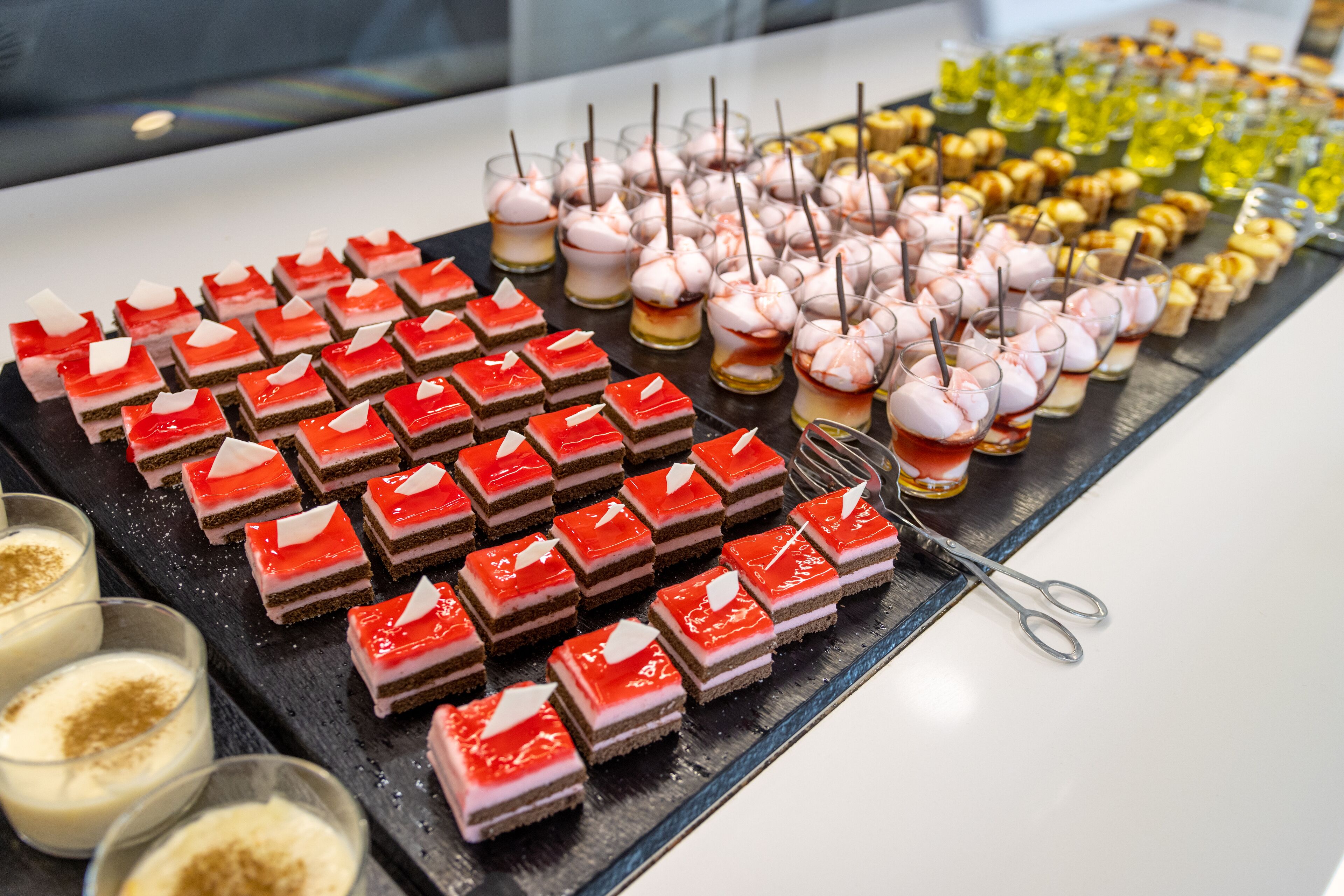 Eine Auswahl an Desserts auf einer Platte, darunter mehrschichtige Kuchen in Rot und Pink, kleine runde Köstlichkeiten und Leckereien in Gelb und Orange.