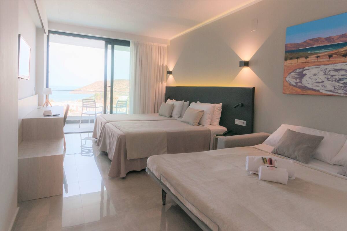 Habitación de hotel con dos camas, un escritorio y una silla. Hay un cuadro en la pared y vista al mar a través de la ventana.