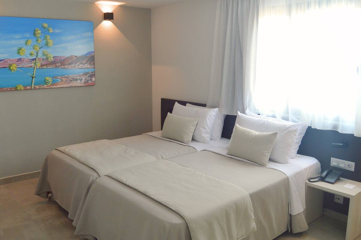 Una cama bien hecha con sábanas blancas, un cuadro de un paisaje en la pared y una mesita de noche con un teléfono.