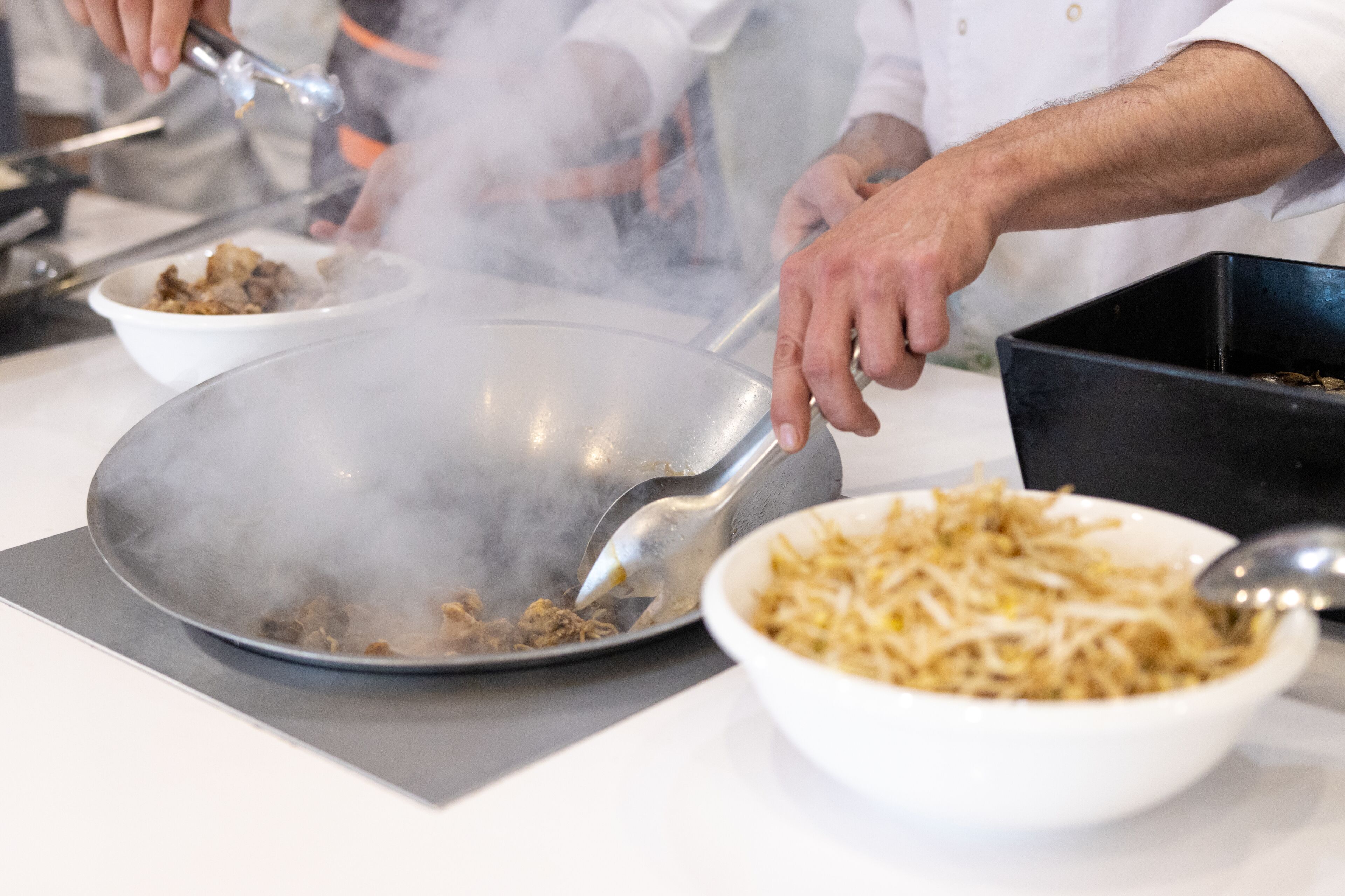 Een kok bereidt eten in een keuken met een grote wok op het fornuis, een schaal noedels en een schaal groenten.