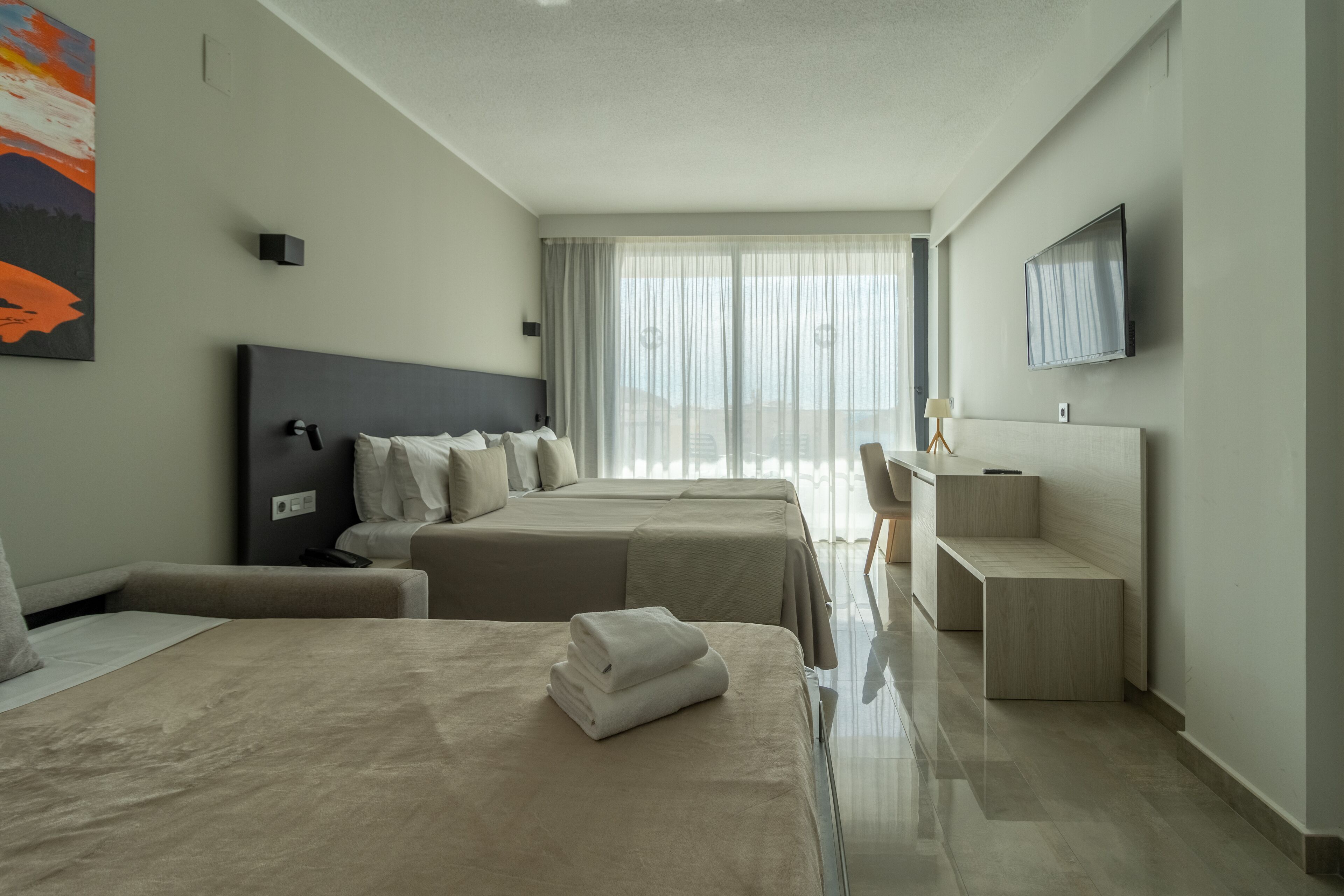 Ein modernes Hotelzimmer mit zwei Betten, einem Flachbildfernseher, einem Schreibtisch und einem großen Fenster mit transparenten Vorhängen.