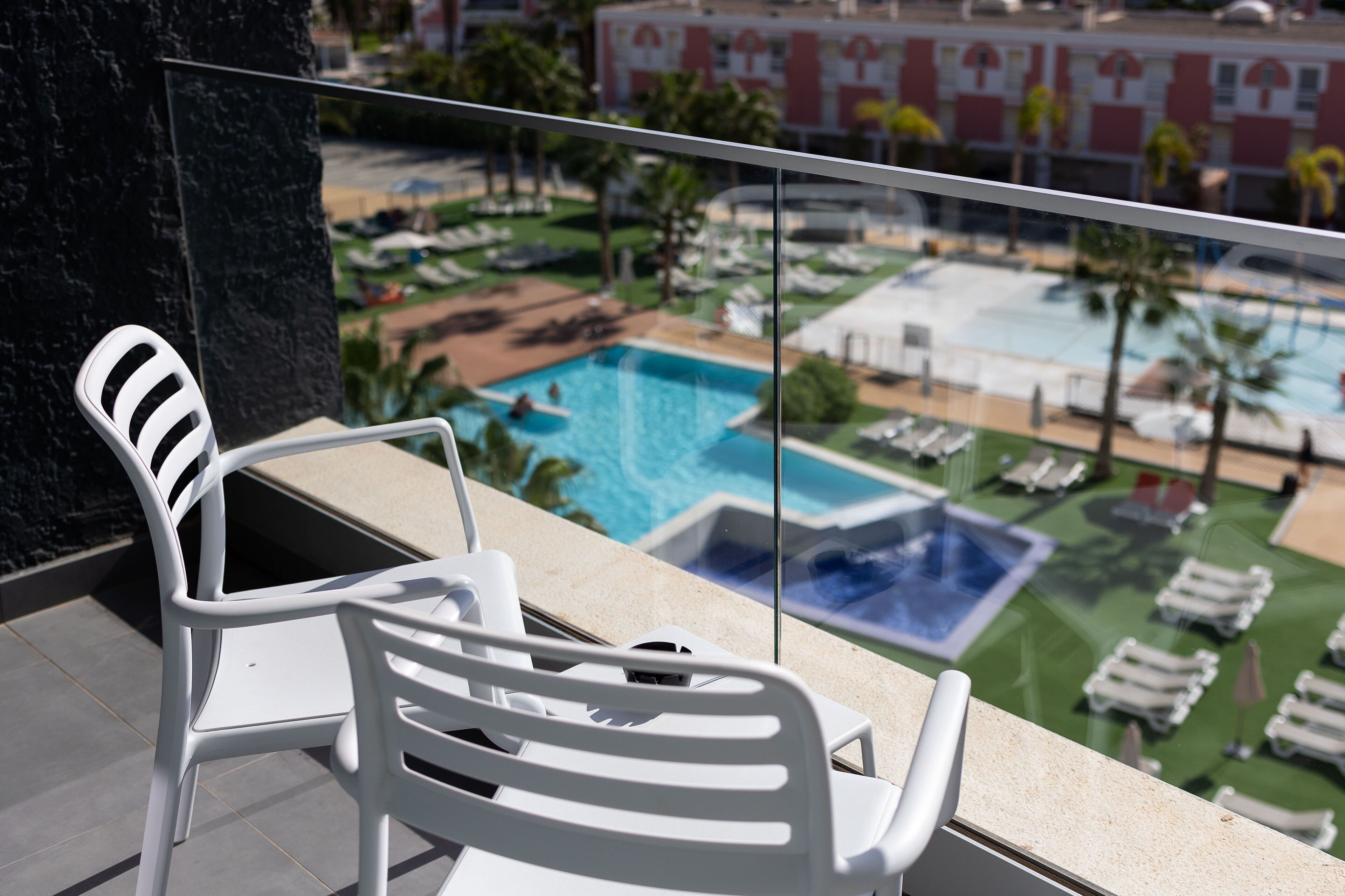 A white chair with a cushioned seat and backrest, positioned on a balcony overlooking a swimming pool area with multiple pools and lounge chairs.