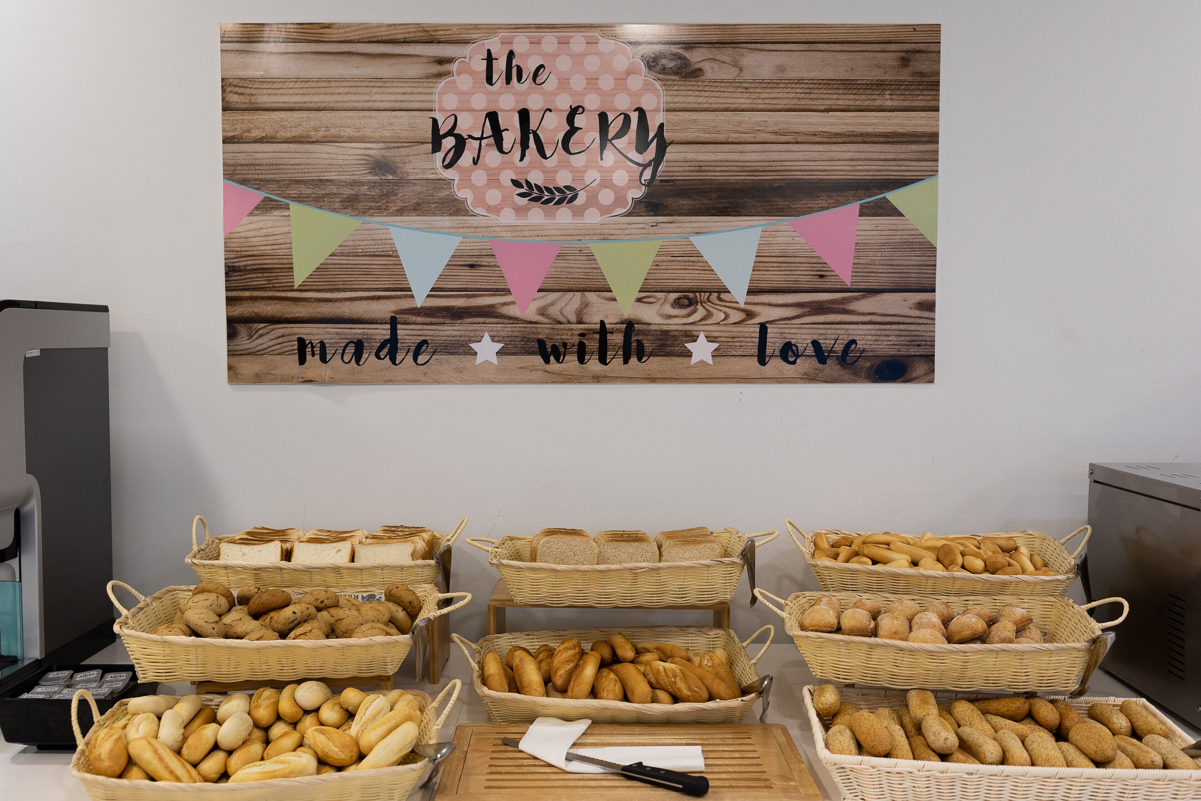 Mostrador de panadería con panes y pasteles variados en cestas de mimbre, un letrero de madera arriba y una tabla de cortar con cuchillo y espátula.