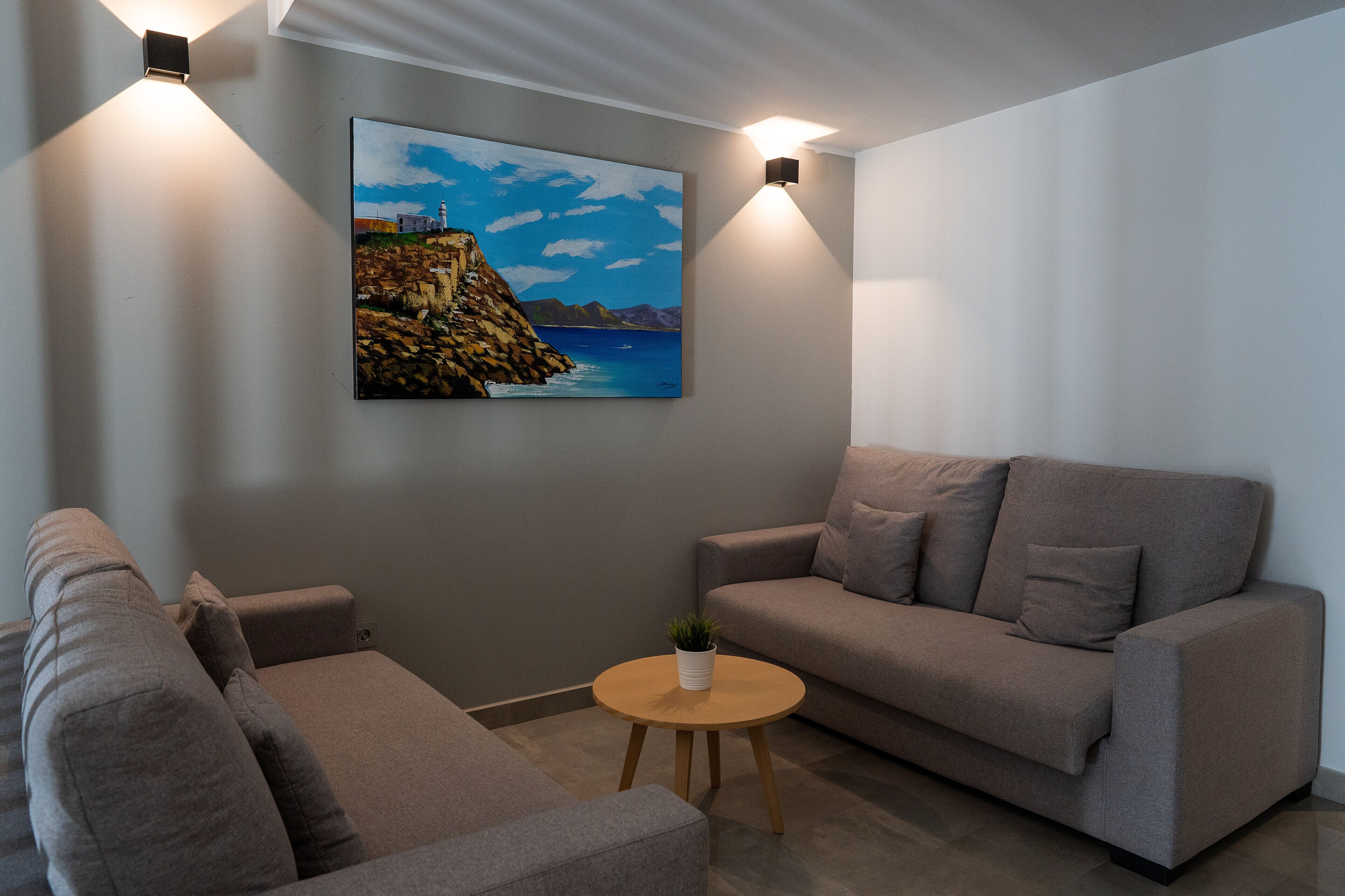 A modern living room with two grey sofas, a small round wooden table with a plant, and a painting of a coastal landscape on the wall.