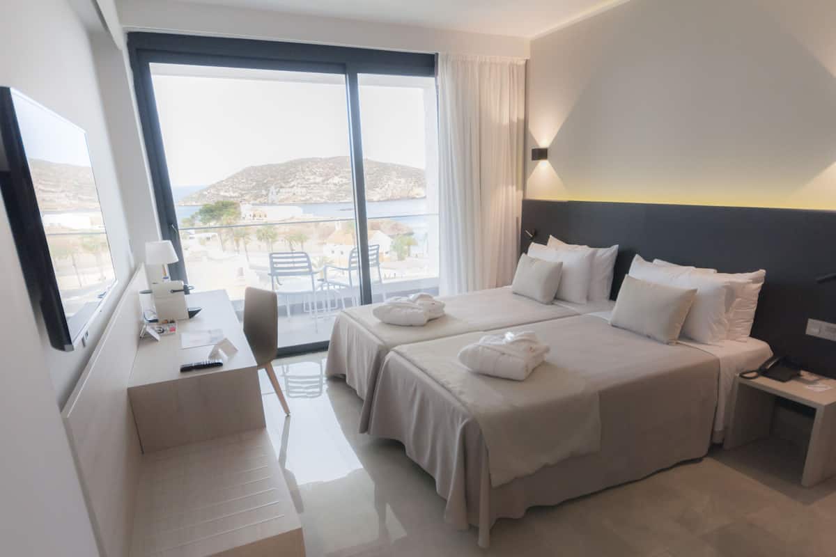 Habitación de hotel con dos camas, un escritorio, una silla y un ventanal con vistas al mar y edificios.