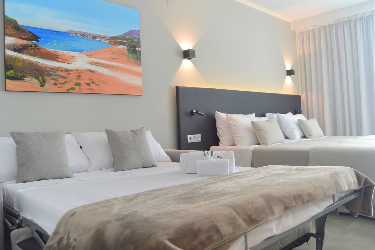 Habitación de hotel con dos camas, un cuadro de playa y una mesita de noche con una lámpara.