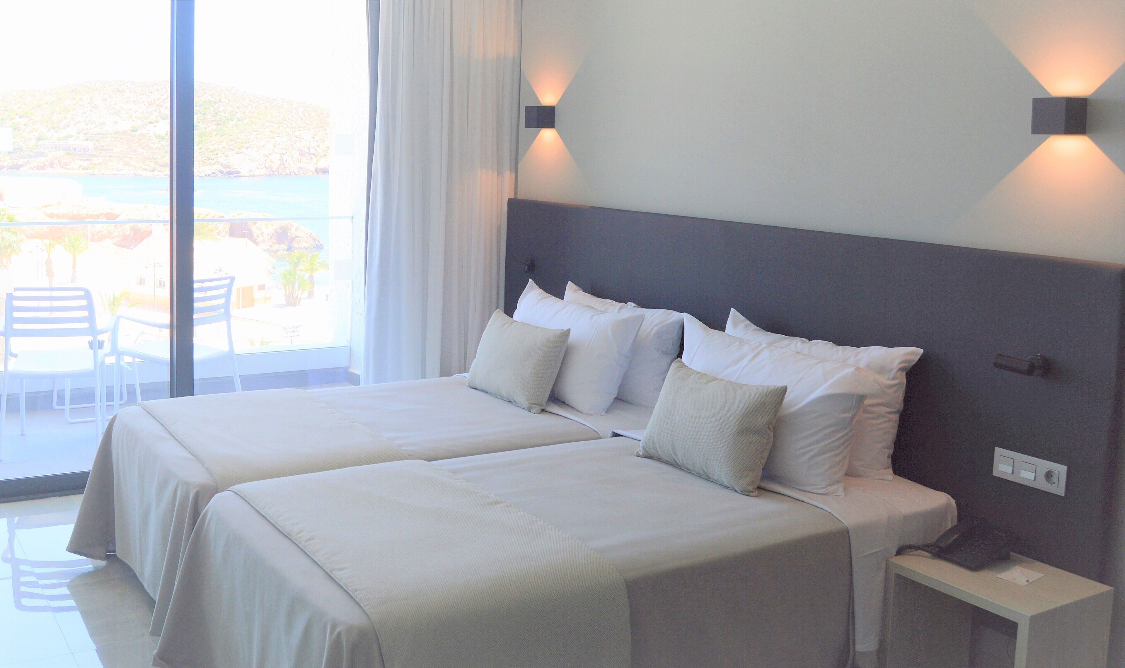 A hotel room with two beds, a nightstand, and a view of the outdoors.