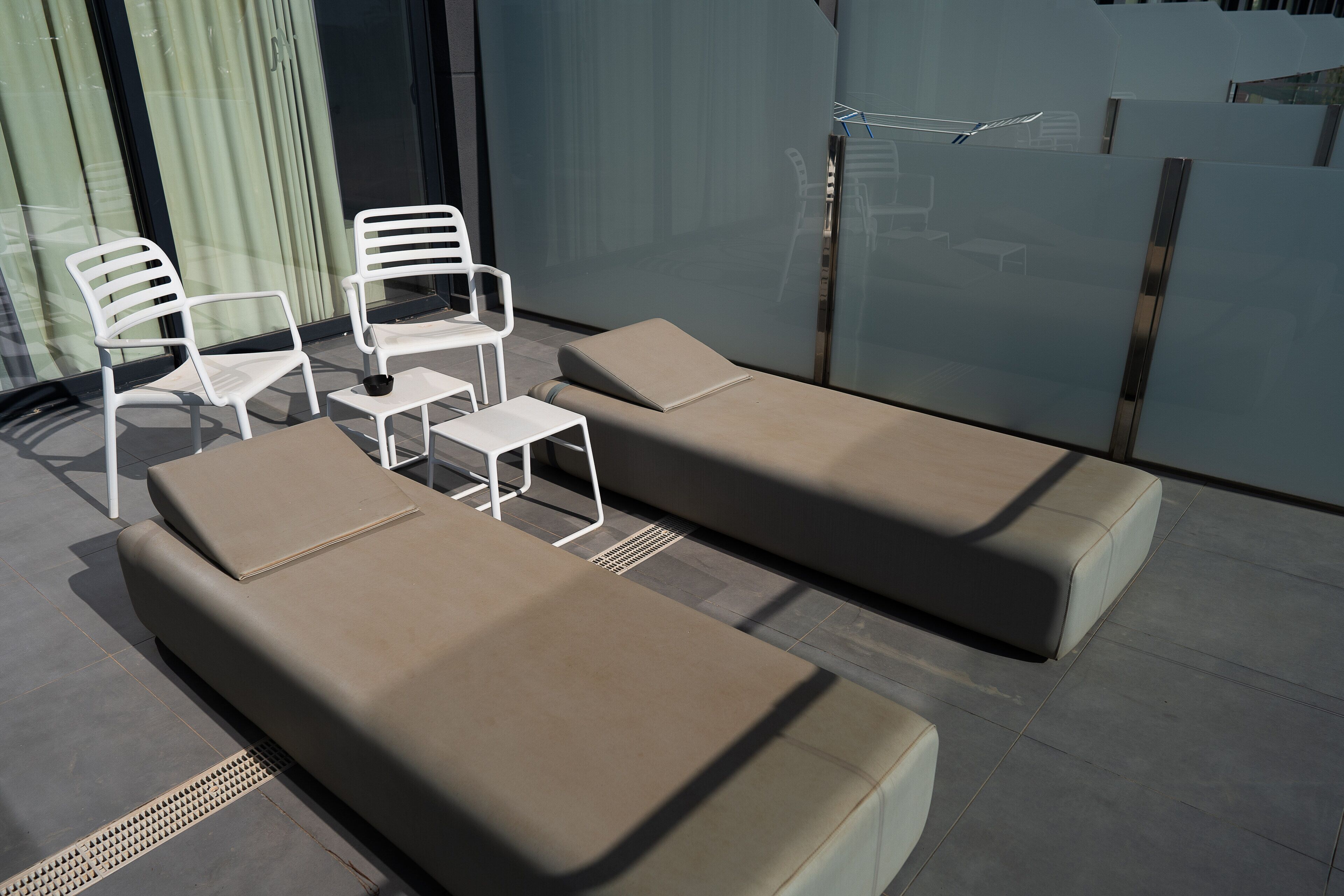 A modern outdoor seating area with two lounge chairs and two white chairs, a small table, and a glass partition.