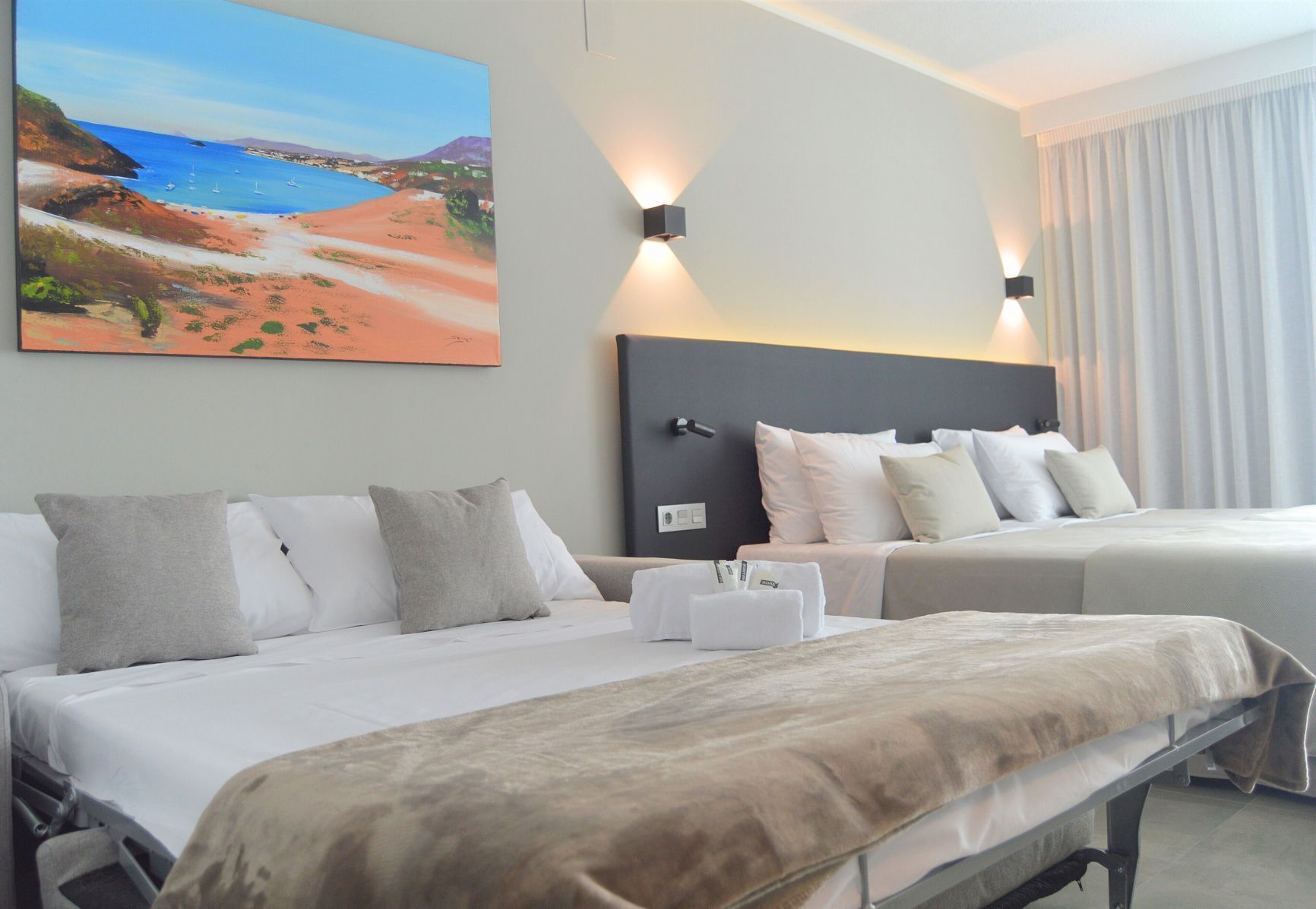 Habitación de hotel con dos camas, un cuadro grande de una playa y una mesita de noche con lámpara.