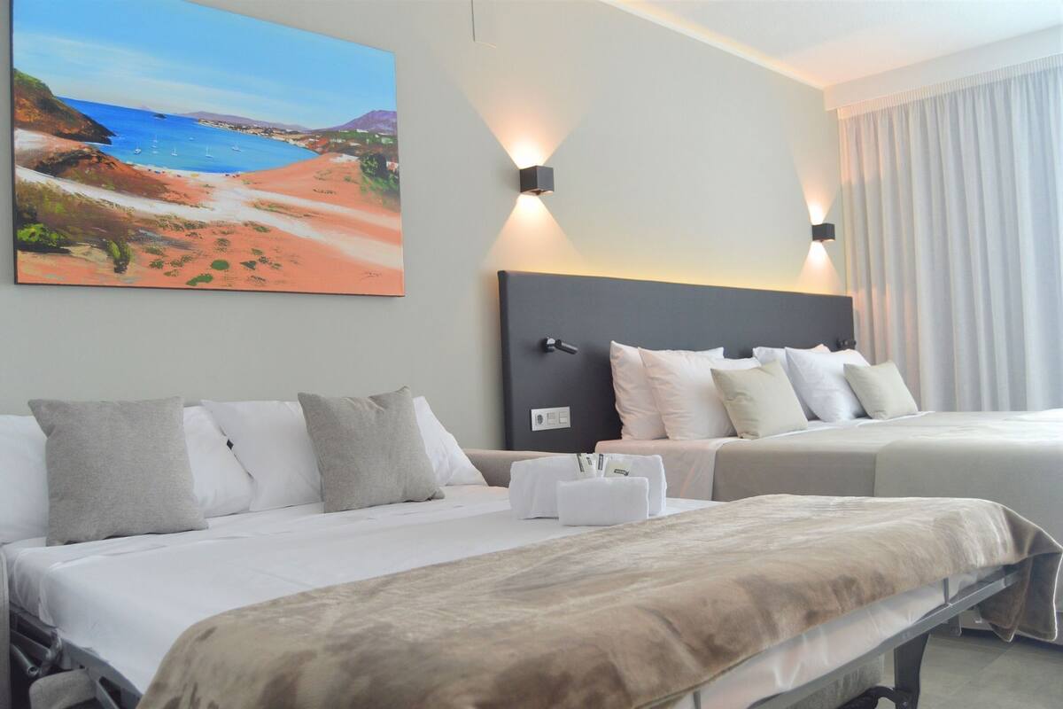 Habitación de hotel con dos camas, un cuadro grande de una playa y una mesita de noche con lámpara.