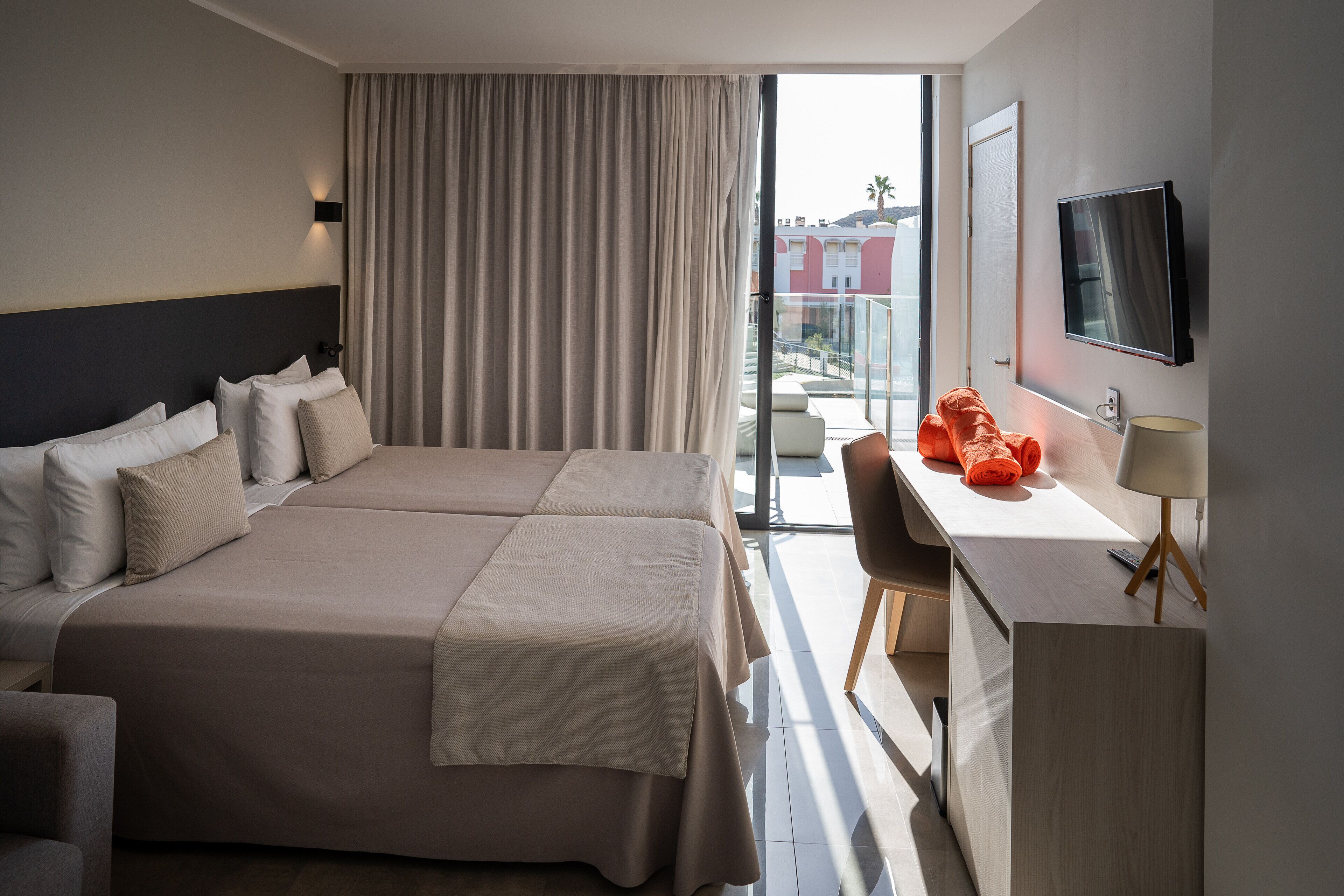 A hotel room with two beds, a desk, a chair, a TV, and a balcony with a view.