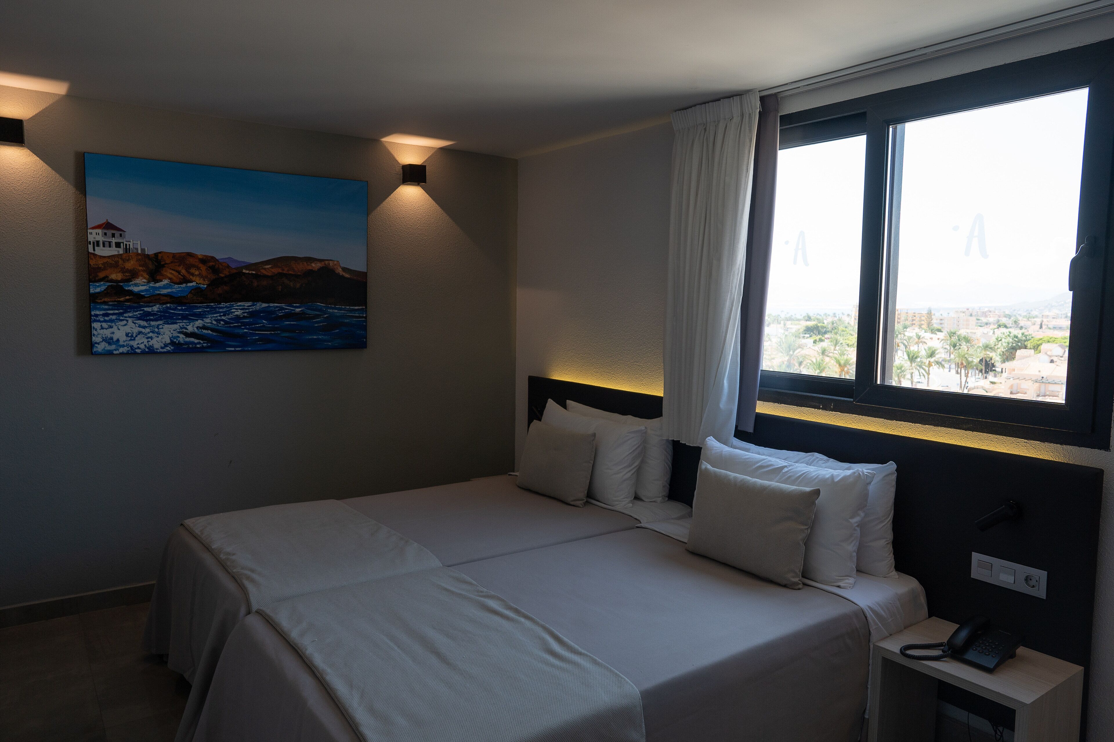 Un cuarto de hotel con cama, mesita de noche, un cuadro de una escena costera y una ventana con vista a la ciudad.