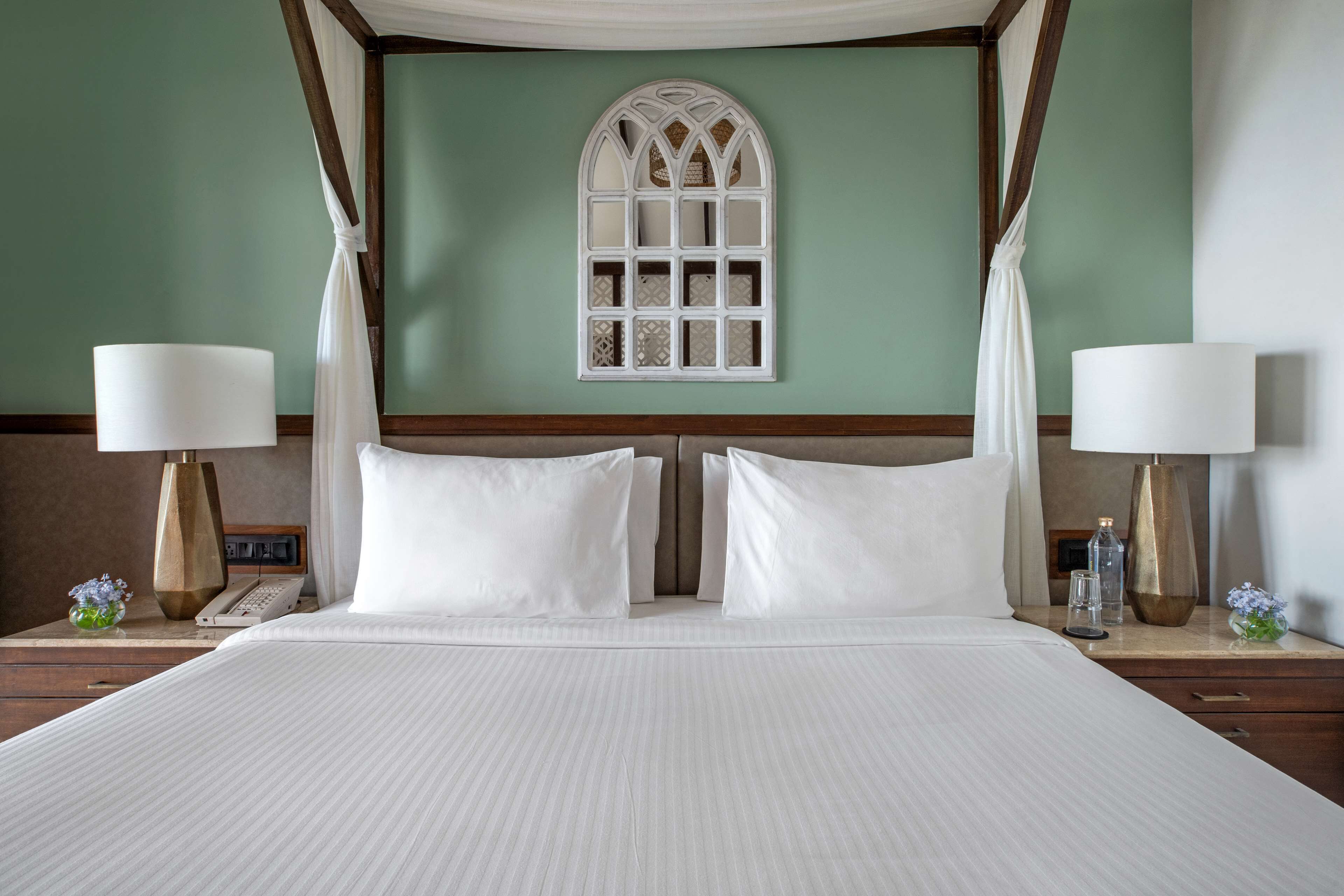 Un lit bien fait, avec du linge de lit blanc, deux tables de chevet avec des lampes, une fenêtre décorative et un mur vert.