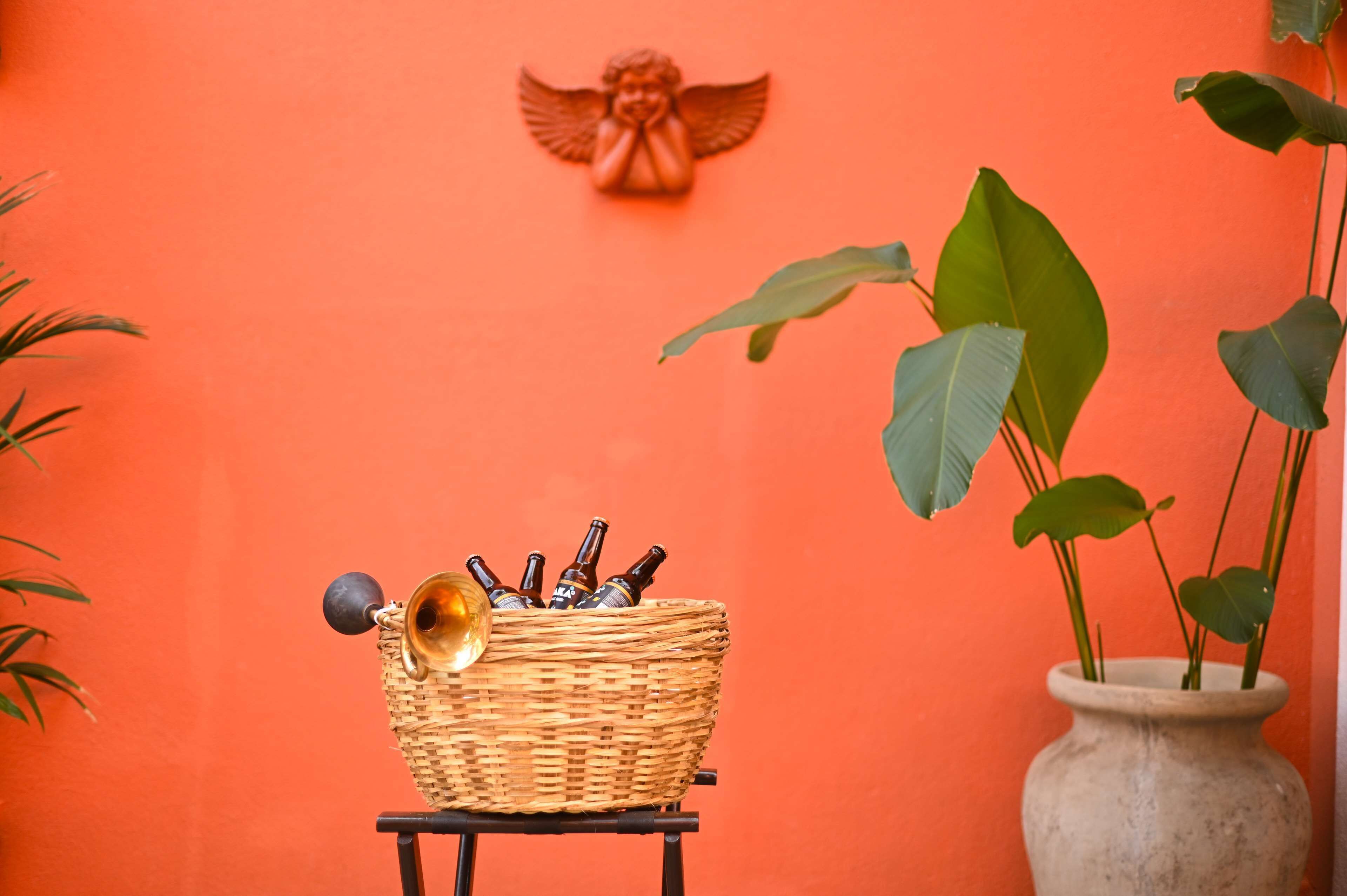 A wicker basket with bottles on a small table against an orange wall, a decorative angel, and a potted plant.