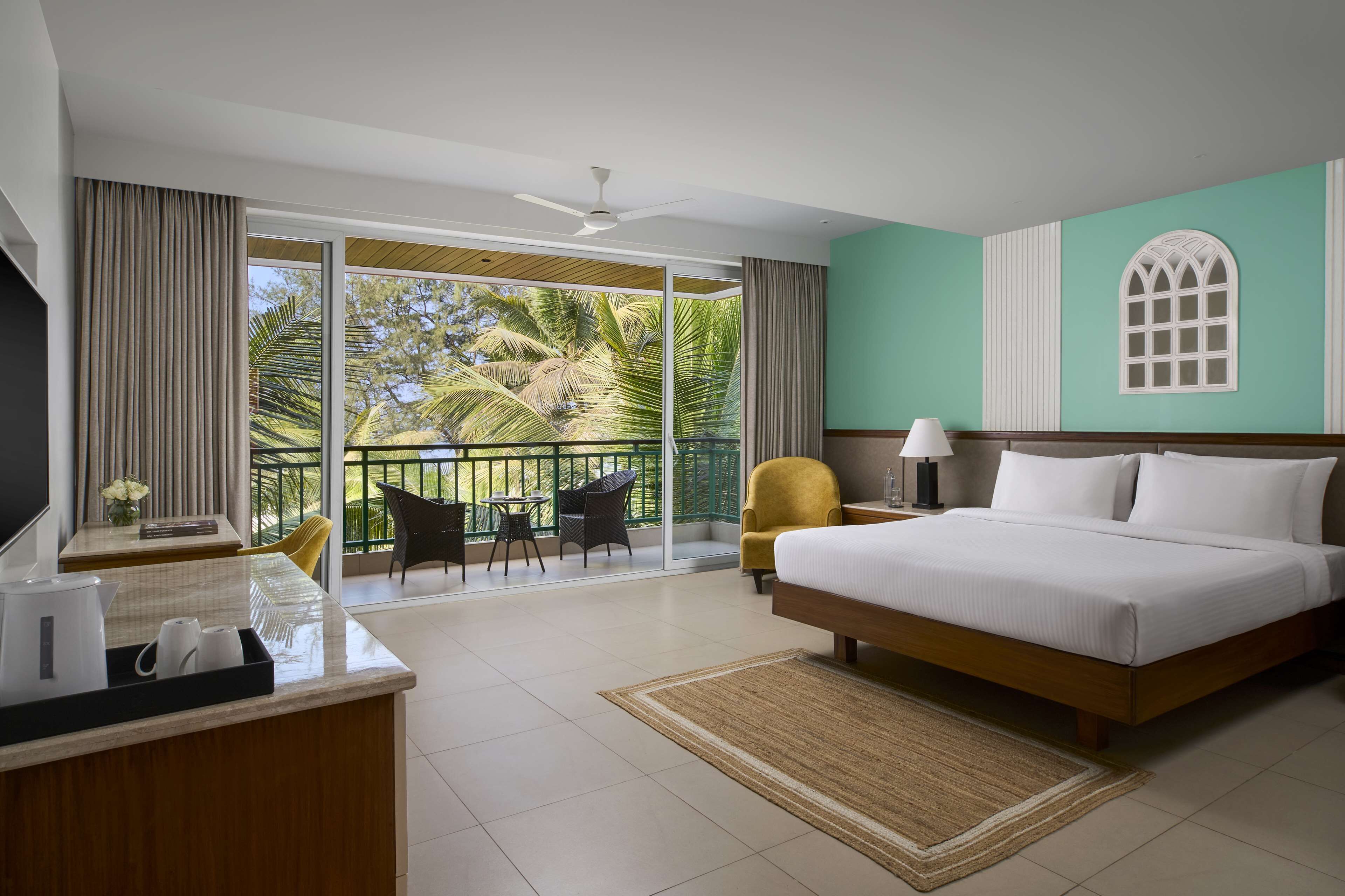 Hotelkamer met een groot bed, een bureau met koffiezetapparaat, een stoel, een balkon met uitzicht op palmbomen en een plafondventilator.