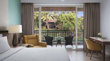 Habitación de hotel con cama, escritorio, dos sillas, lámpara y balcón con vista a palmeras y un edificio.