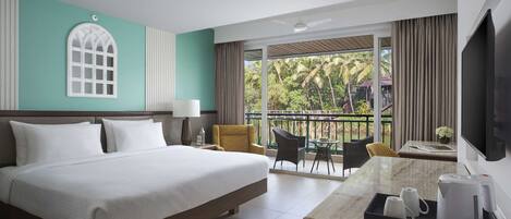 A hotel room with a large bed, a desk with a coffee maker, a TV, and a balcony with a view of palm trees.