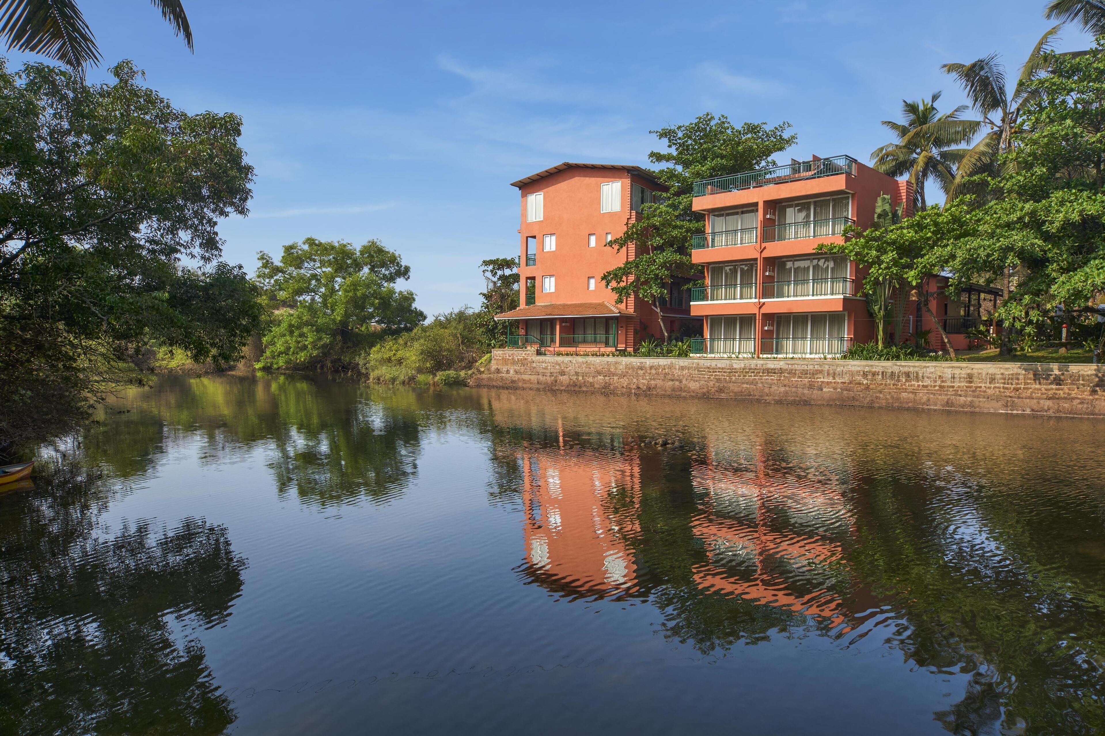A multi-story building with balconies, situated beside a calm river, surrounded by lush greenery.