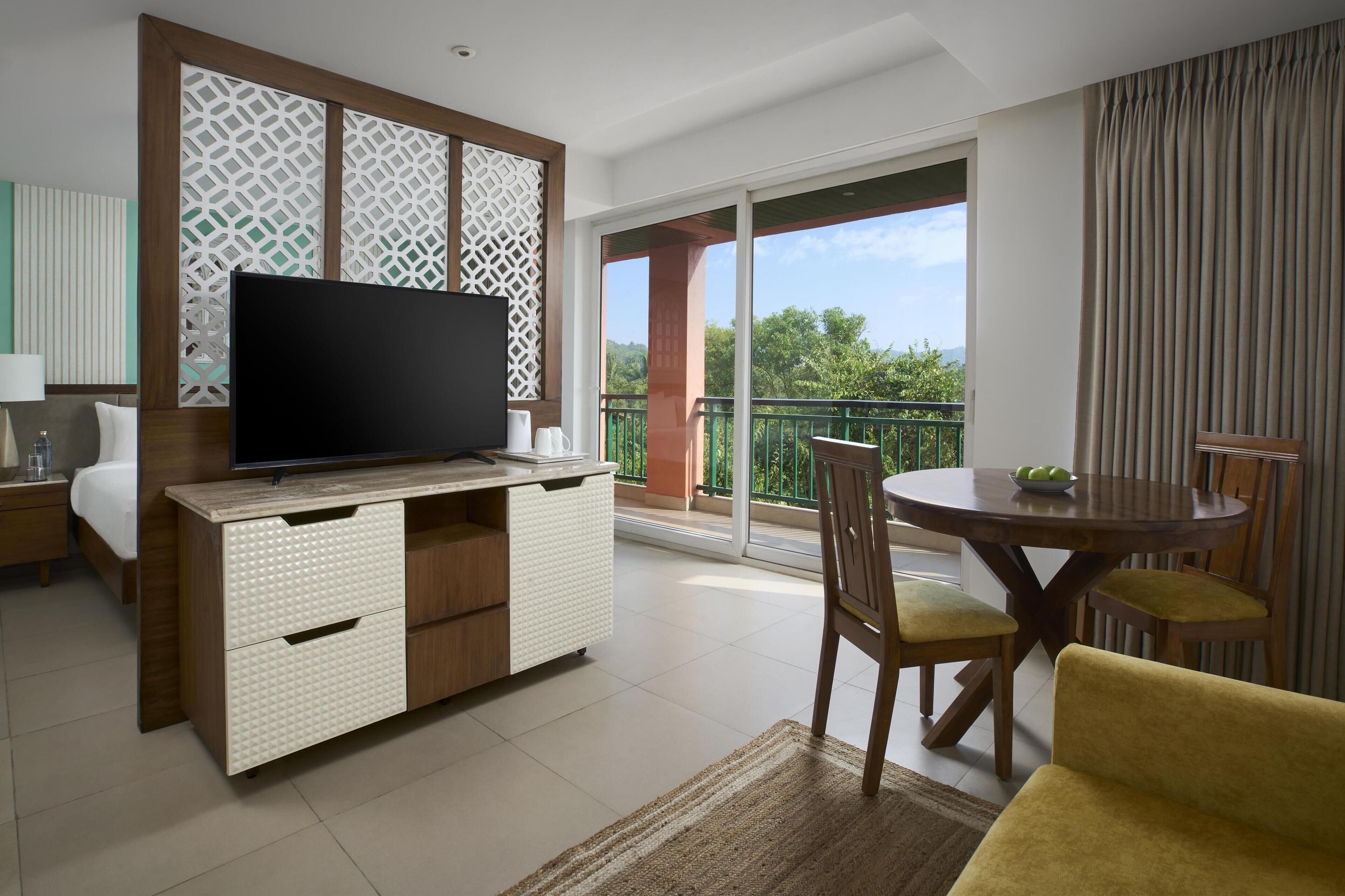 A modern hotel room with a flat-screen TV, a wooden dining table with chairs, a sofa, and a balcony with a view of greenery.