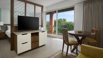 A modern hotel room with a flat-screen TV, a wooden dining table with chairs, a sofa, and a balcony with a view of greenery.