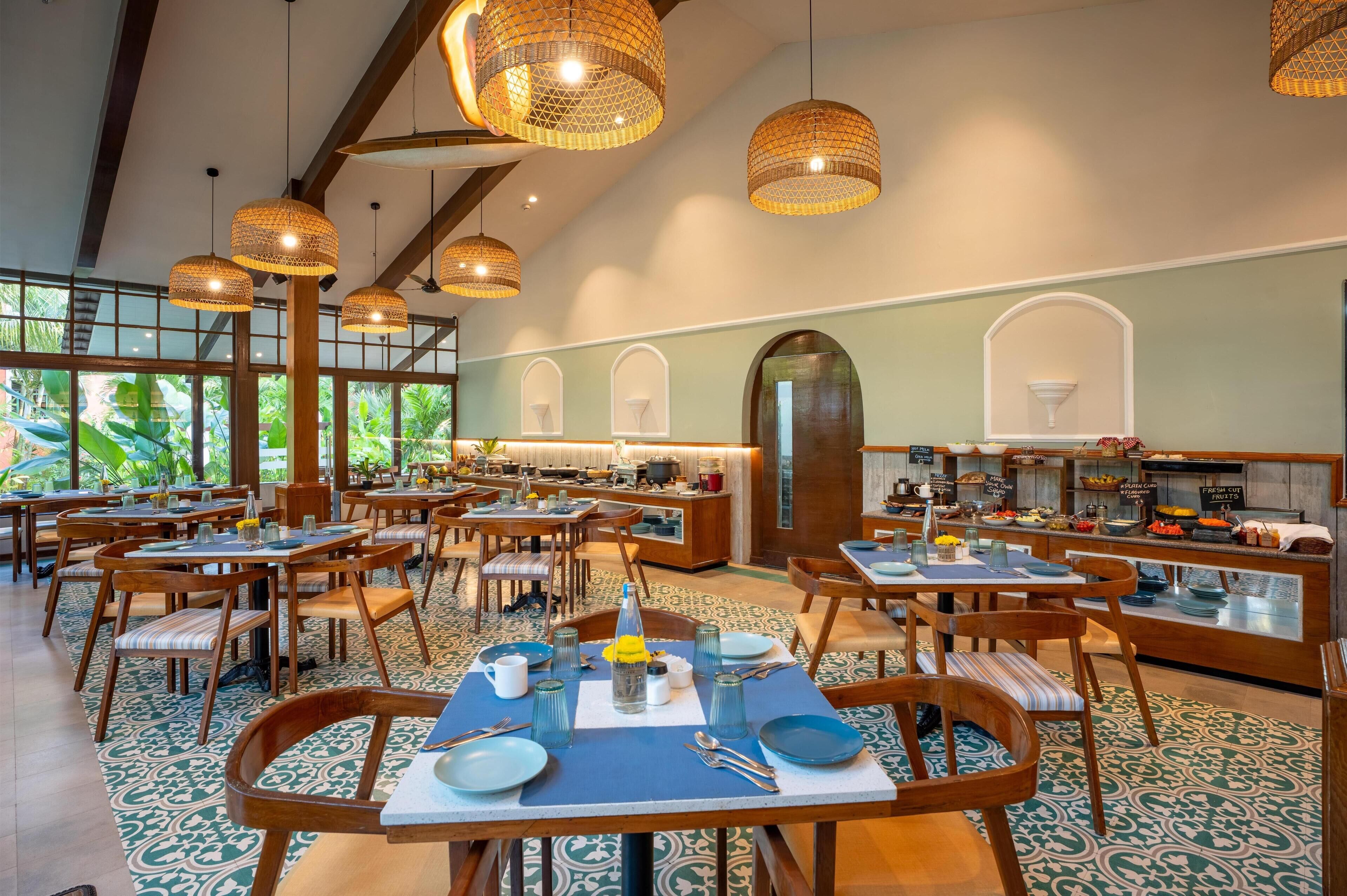 A spacious dining area with multiple tables and chairs, large windows with a view of greenery, and hanging wicker lamps.