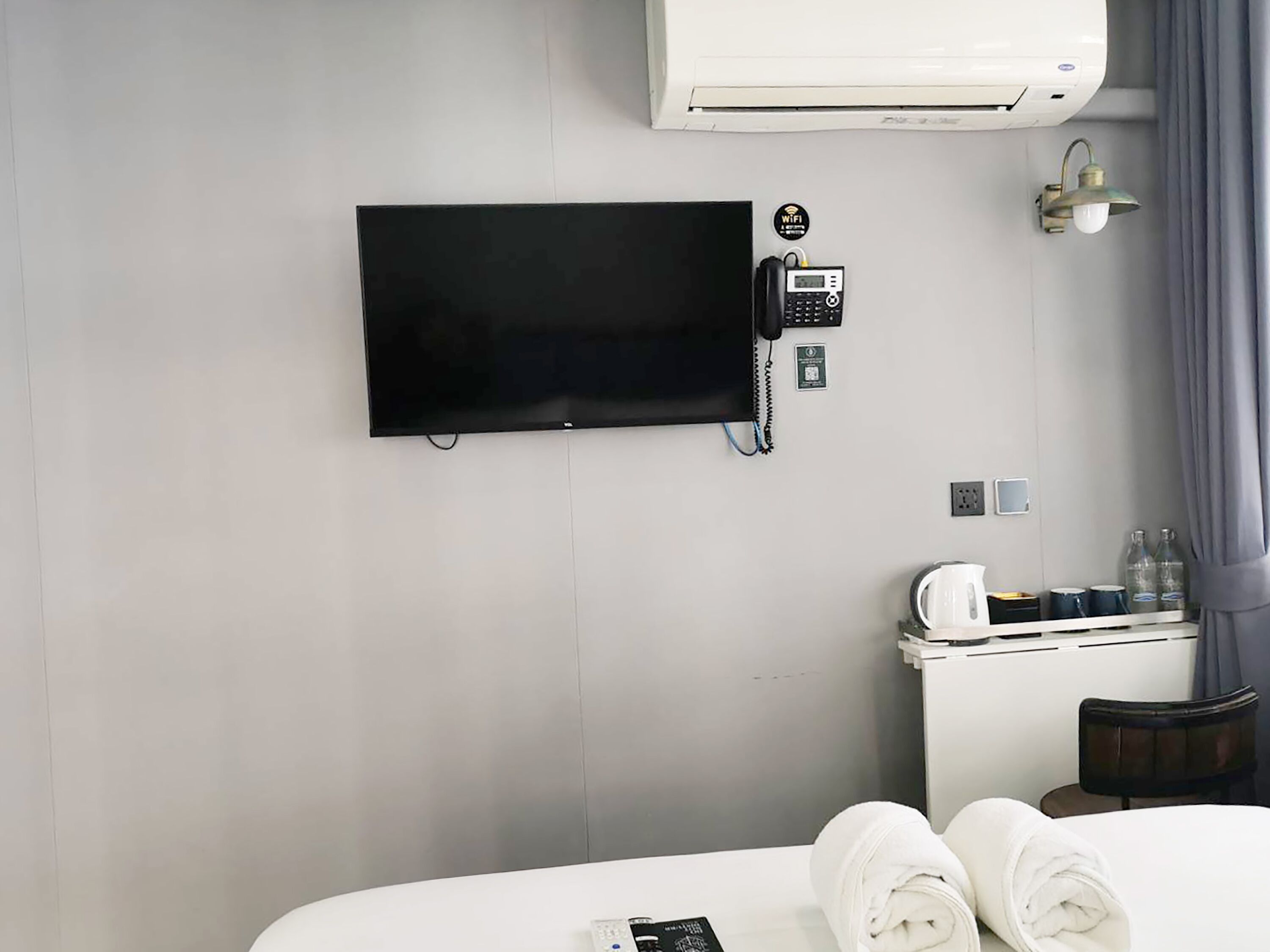 Camera d'albergo con televisore a schermo piatto montato a parete, un minibar, un letto con asciugamani arrotolati e un condizionatore d'aria a parete.