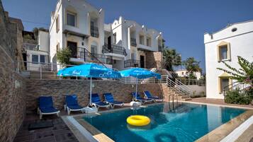 A swimming pool with lounge chairs and umbrellas, surrounded by buildings with balconies and stone walls.