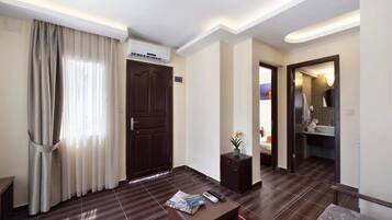 A hotel room with a dark wood floor, a large dark wood door, a small table with a remote control, a potted plant, and a bathroom visible through an open door.